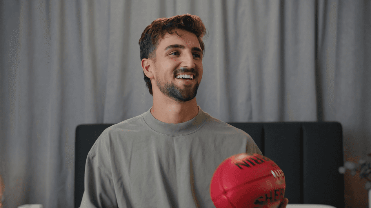 A young man with short hair wearing a light-colored t-shirt sits in an indoor setting, holding a red boxing glove