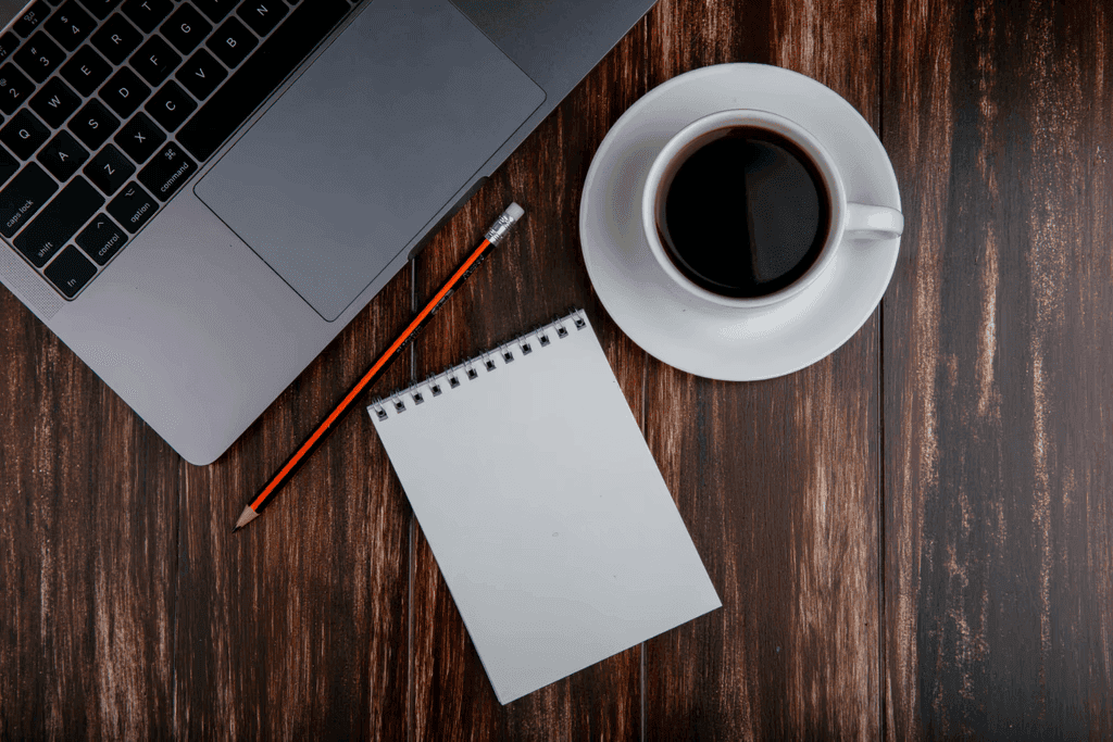Laptop, blank notepad, pencil, and a cup of coffee on a wooden desk