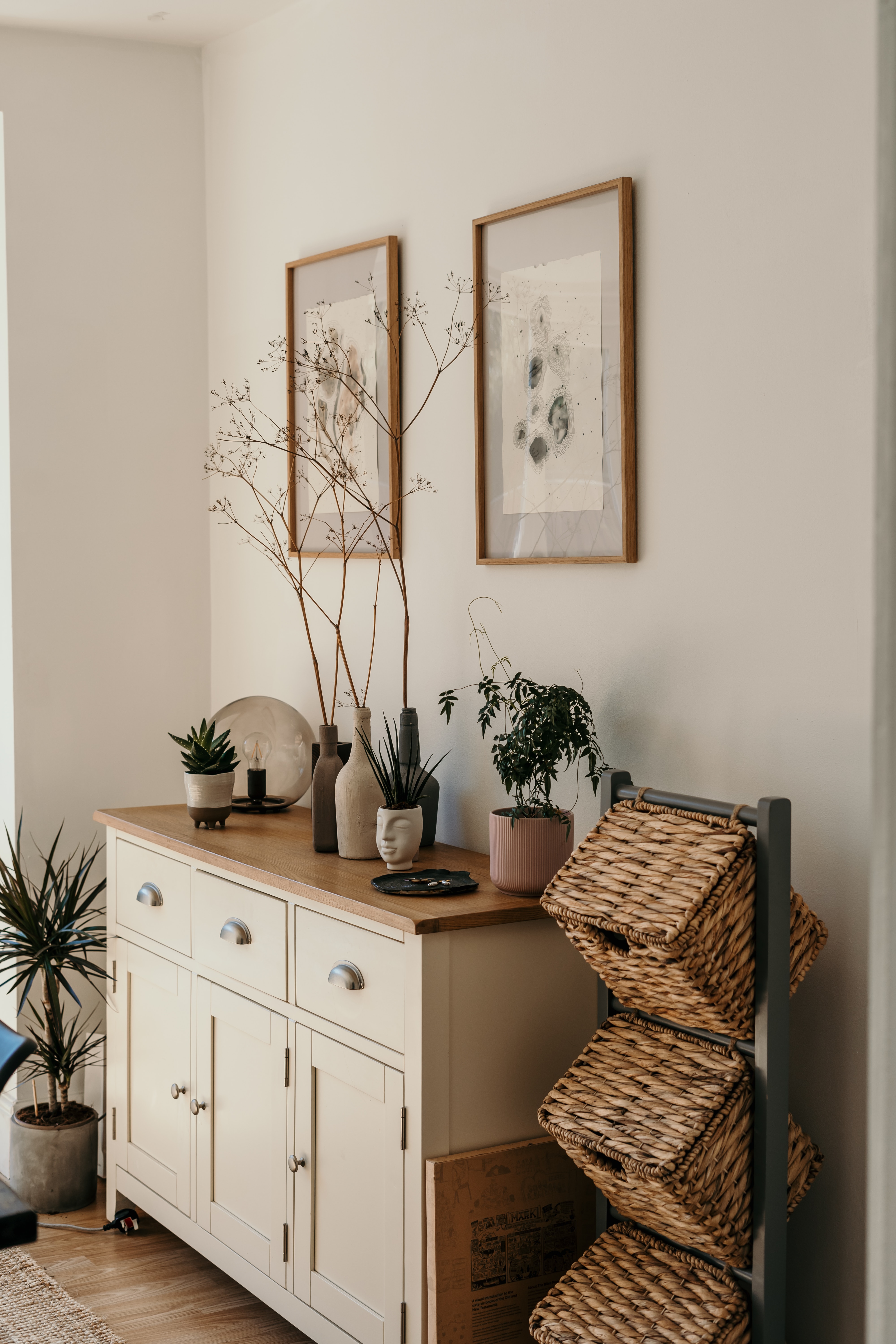 Image of home interior design showing a dresser with a wooden countertop, some braided baskets and 2 frames with some abstract art hanging on the wall.