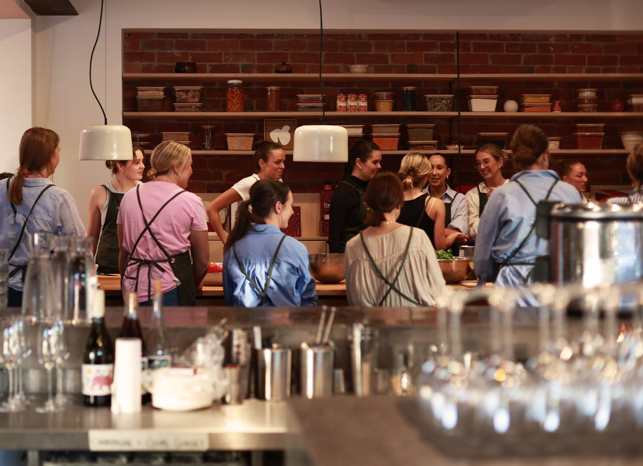 Group of guests wearing aprons standing at a kitchen bench in front of shelves stocked with pantry jars and containers