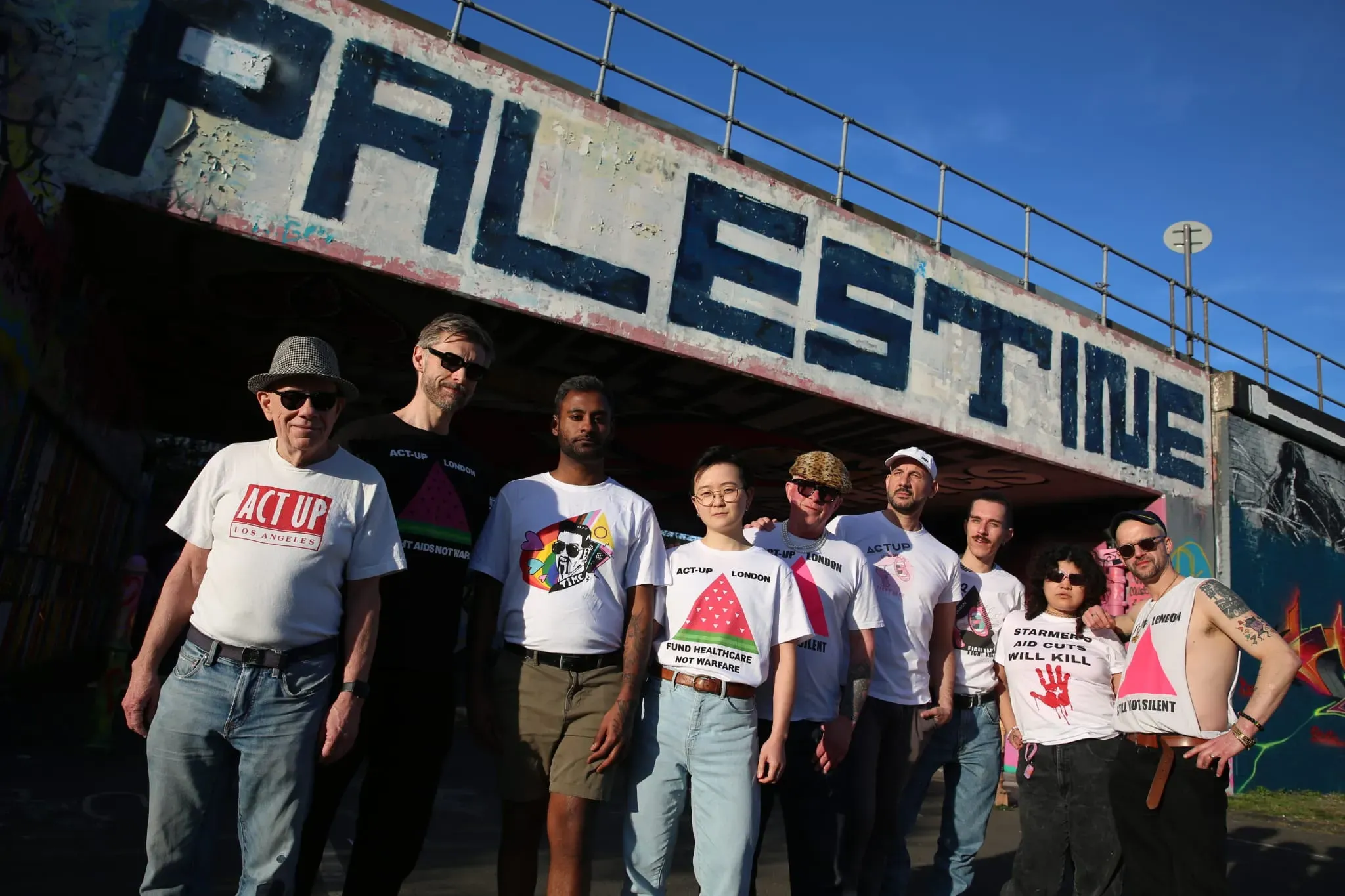 A group of Act Up activists wearing a variety of Act Up t-shirts stand in front of a bridge which has the word Palestine written on it in black paint.