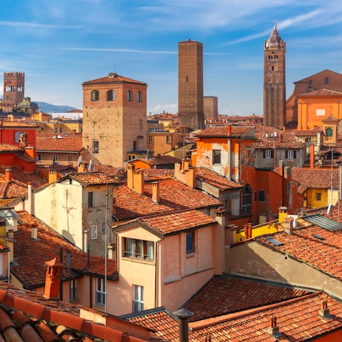 Un panorama urbano di Bologna, Italia, che mostra edifici dai tetti rossi, torri e un cielo azzurro limpido sullo sfondo.