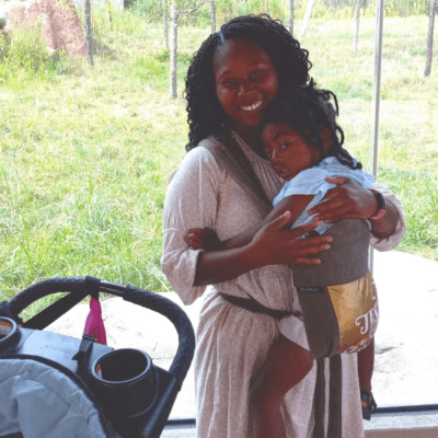 Alt tag: Portrait of Itarah Godbolt smiling while holding a young child in a front carrier, standing near a window with greenery visible outside.