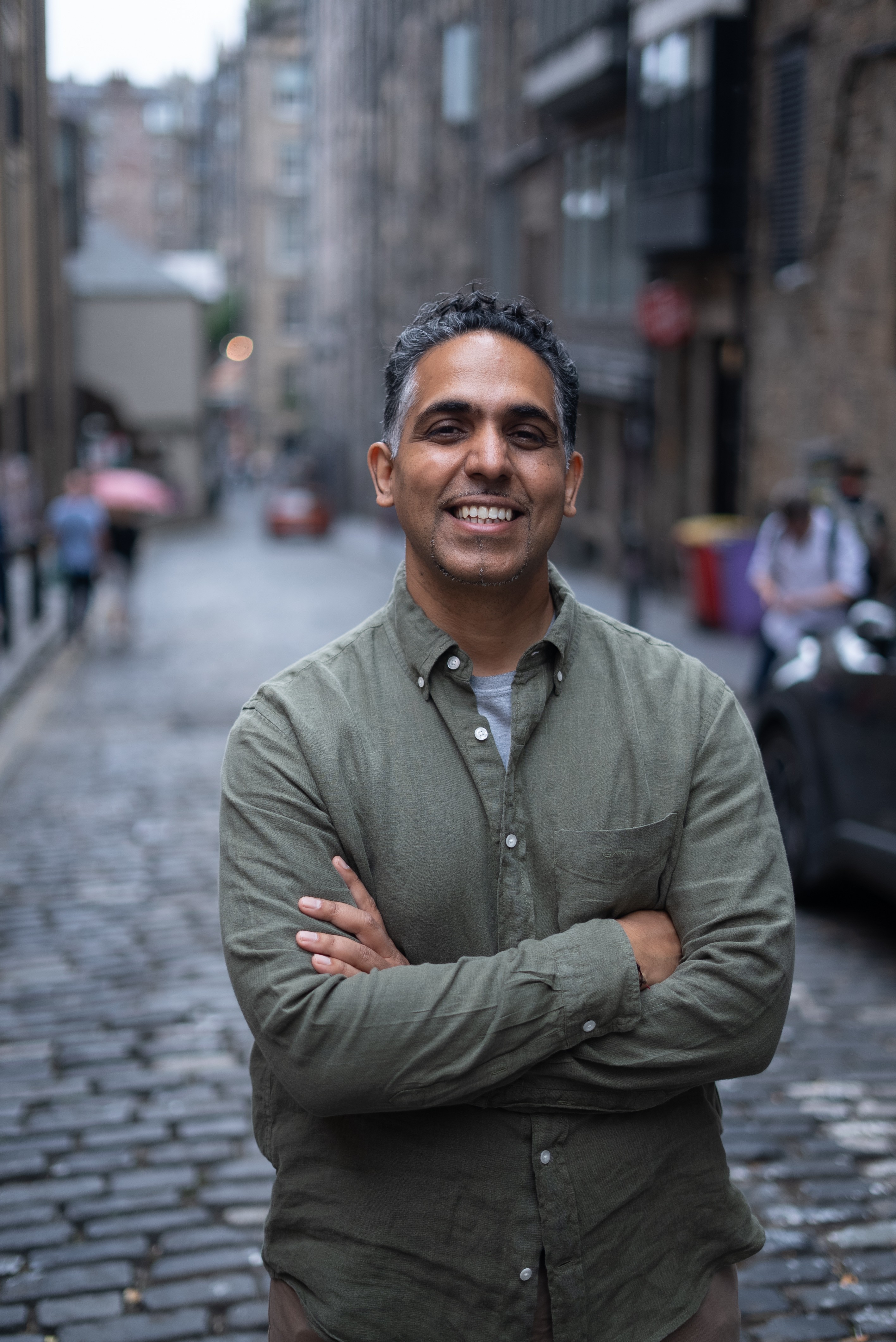 A smiling man with short hair stands with arms crossed, wearing a dark shirt against a neutral background.