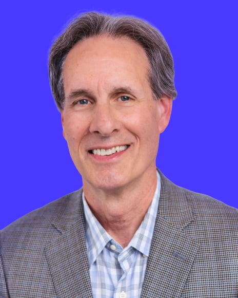 Rob Rash, Director, professional headshot wearing a gray blazer and light blue checkered shirt, smiling at the camera against a vibrant blue background.