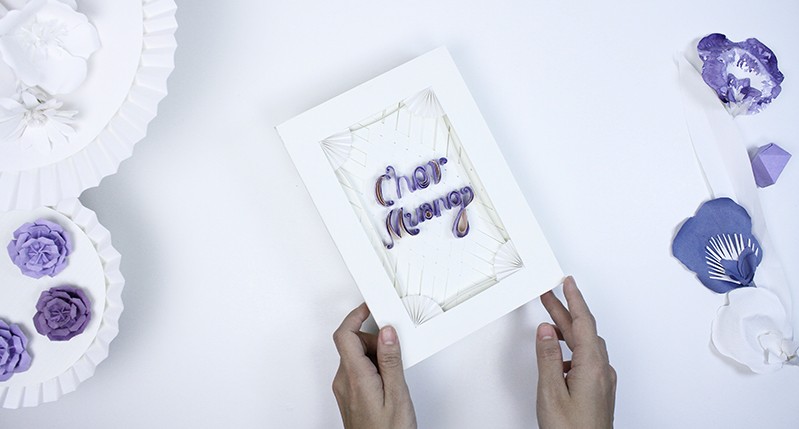 A photo of two hands holding a white box with some purple writing on it. Also there are some white and purple floral arrangements near.