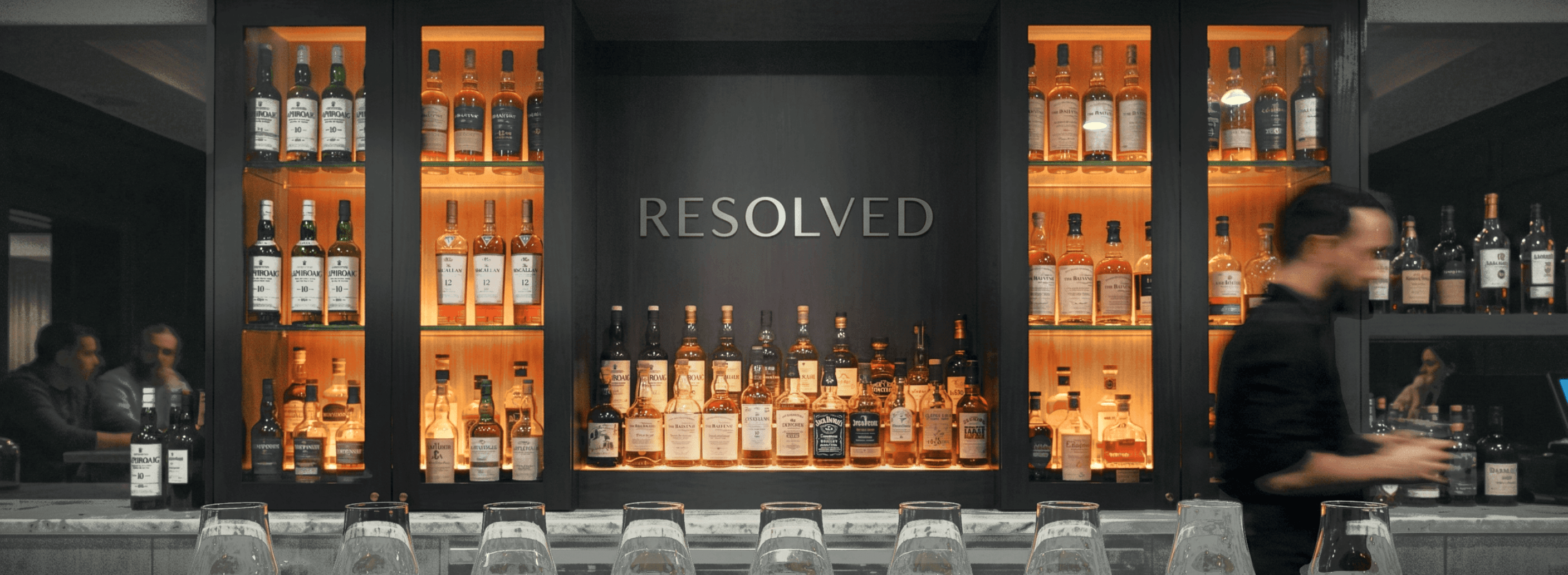 A sophisticated bar interior featuring shelves lined with various whiskey and liquor bottles, subtly illuminated against a dark backdrop with the word "RESOLVED" prominently displayed.