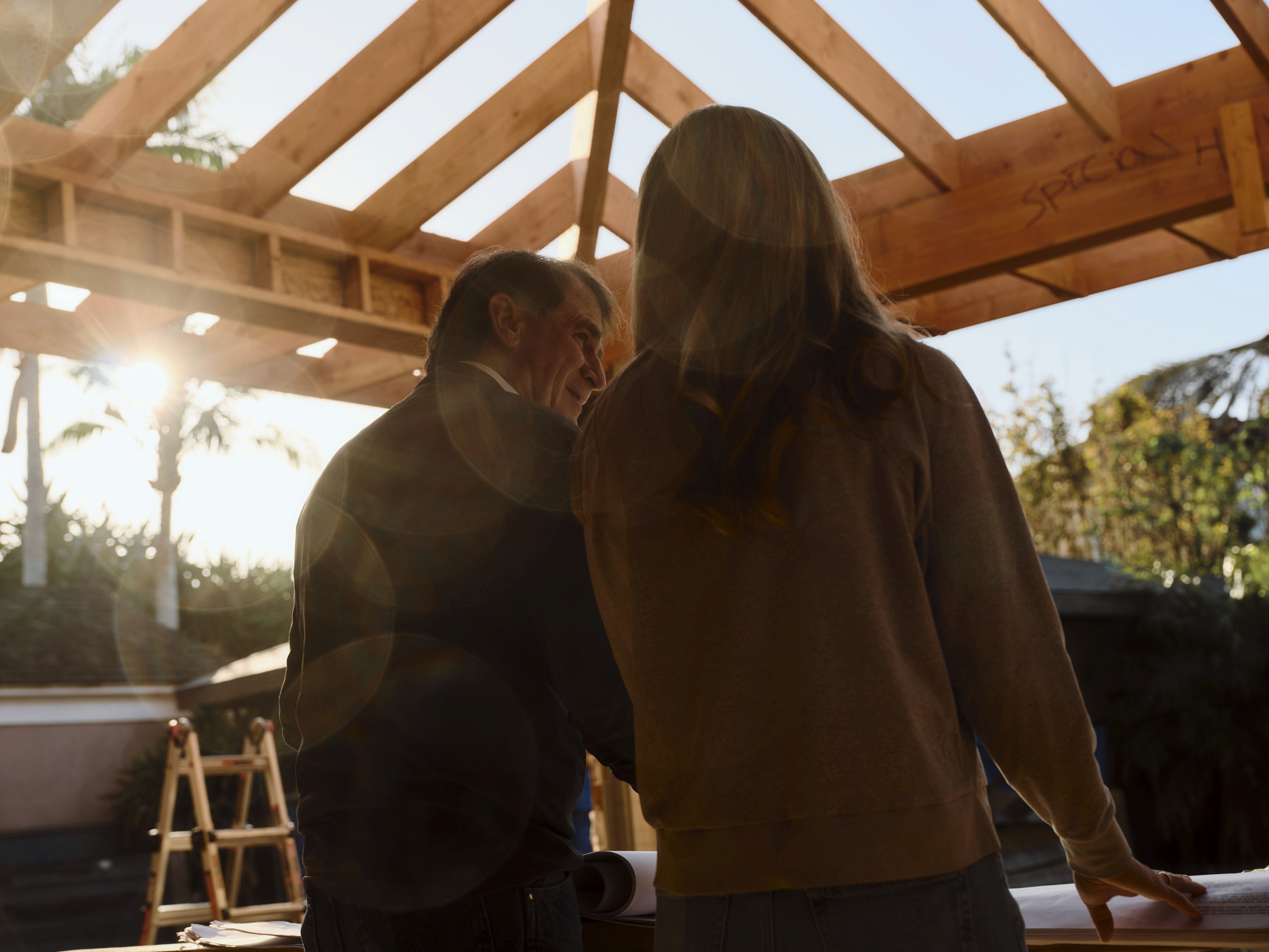 Two people review architectural plans in a sun-drenched, unfinished timber roof frame structure. A man looks towards a woman while they stand over documents.