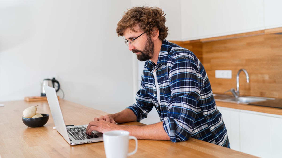 A man standing in the kitchen and working on his computer while drinking a coffee 
