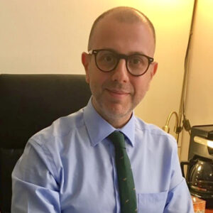 A young person with curly hair smiles at the camera while sitting in front of a laptop against a dark background.