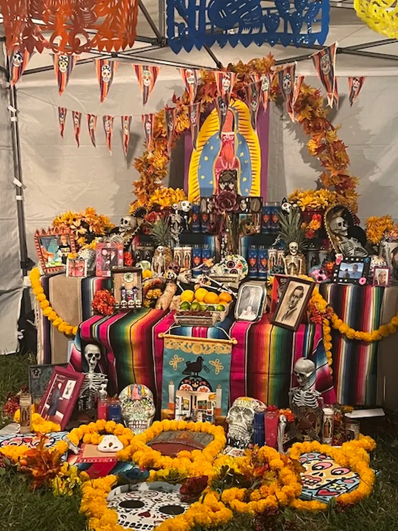 An elaborately decorated Día de los Muertos altar in the Village of the Arts, Bradenton, FL, featuring vibrant marigold flowers, colorful papel picado, and various adorned skulls and skeletons.