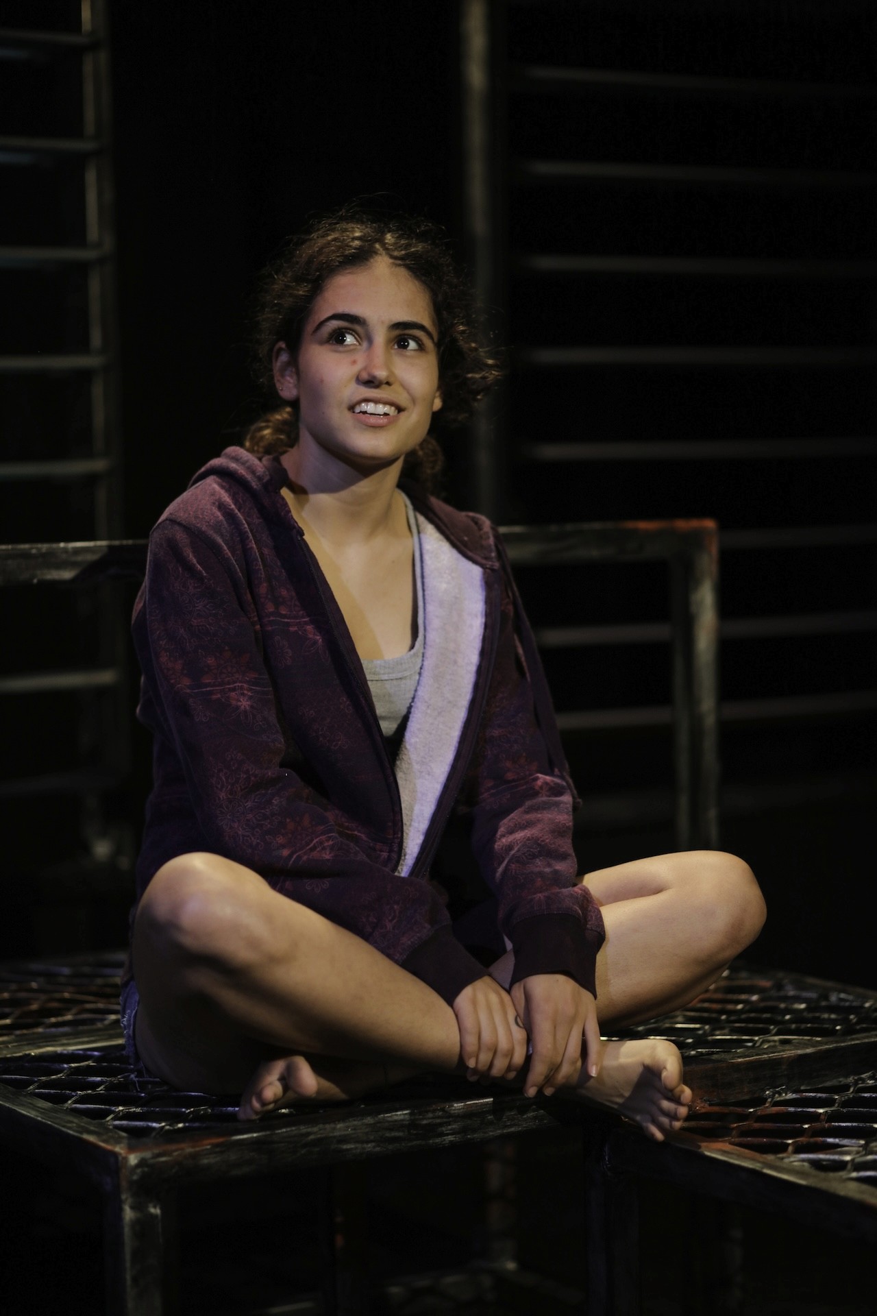 A girl sits cross-legged on a metal platform, wearing a maroon hoodie and shorts. She looks upwards with an open, hopeful expression against a dark industrial backdrop.