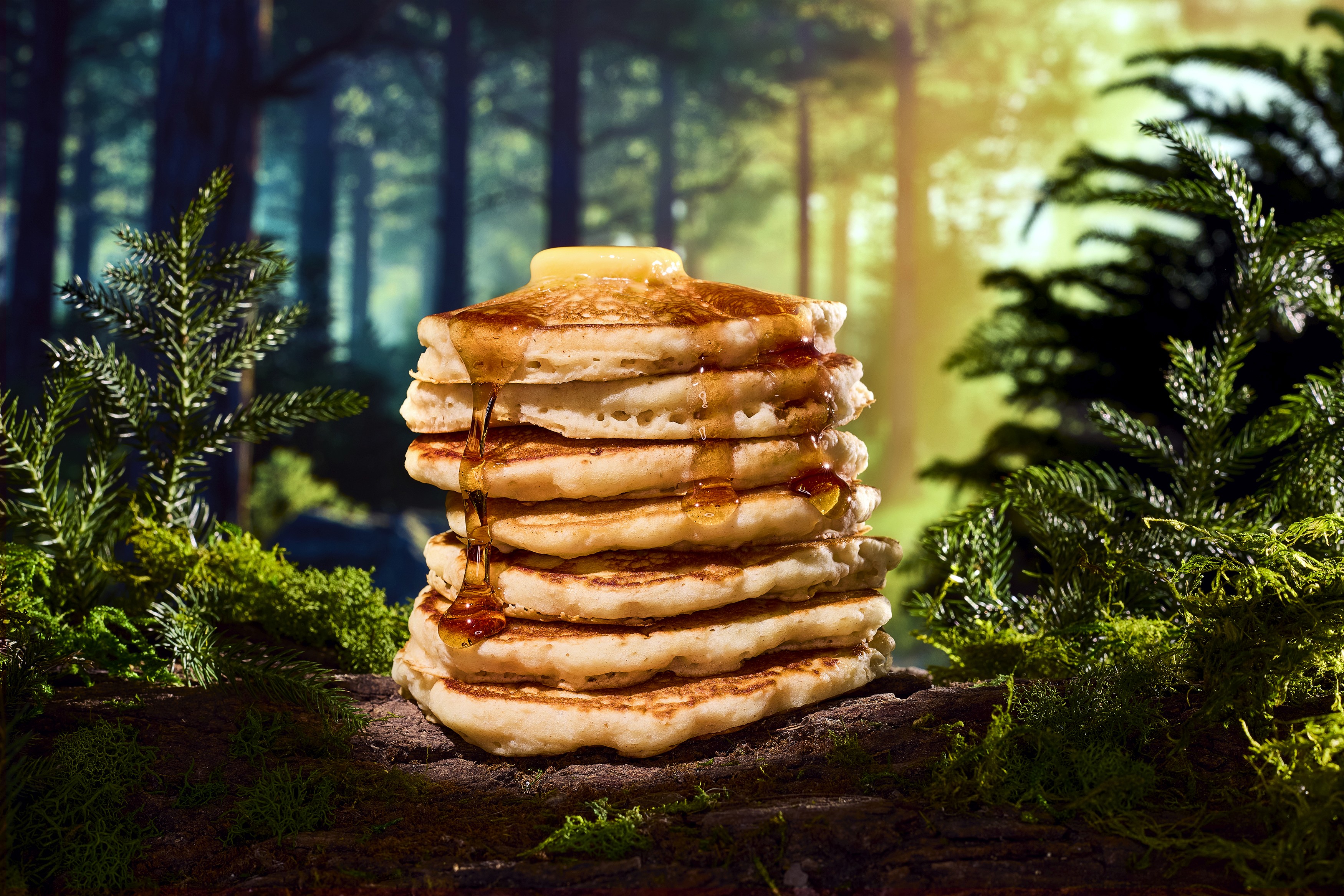 A tall stack of 7 fluffy pancakes sitting on a fallen log amongst the ferns with maple syrup pouring down the sides and a pat of butter melting on top, with morning sunlight streaming through the trees.