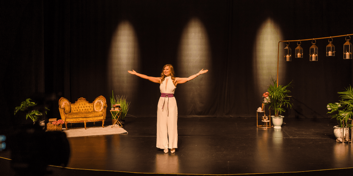 Woman presenting on a decorated theater stage with her arms extended under warm spotlights.