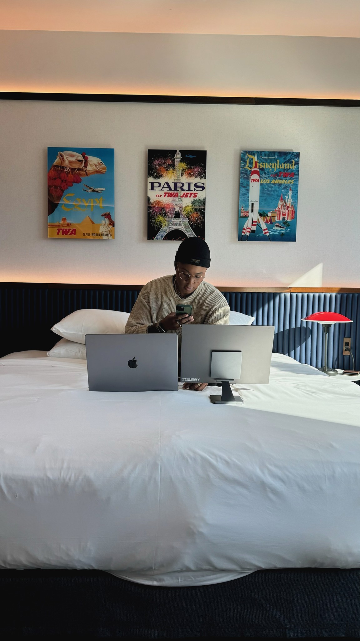 A well-decorated hotel room with a bed, decorative artwork on the walls, and Ashleigh Warren sitting on a hotel bed working on a laptop.