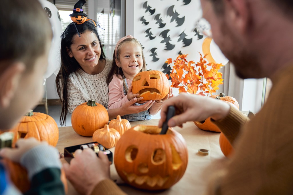 A family enjoys a fun pumpkin carving session at home, with a mother and daughter smiling proudly at their carved pumpkin, surrounded by other pumpkins and festive Halloween decorations like bats on the wall and autumn leaves on the table.