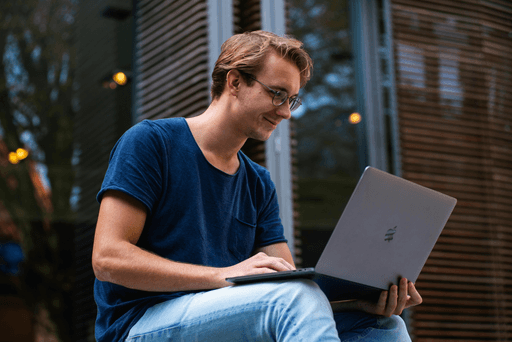 A young man sits outdoors, focused on his laptop while surrounded by greenery and a modern building.