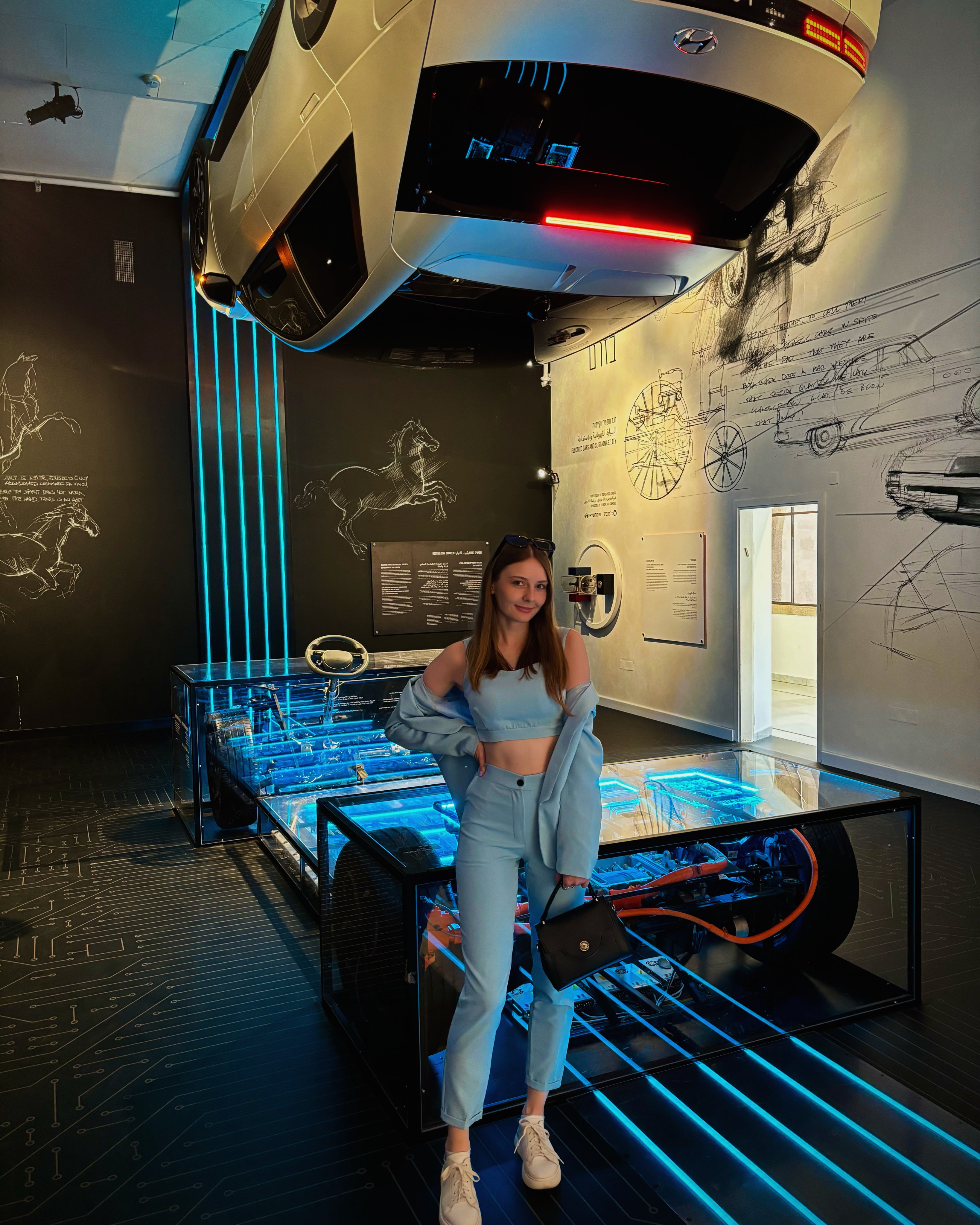 Liana Tudakova — founder of Luna UI Design Studio, standing in a futuristic automotive design exhibition, surrounded by neon light and engineering sketches, reflecting her passion for innovation, systems, and modern aesthetics.