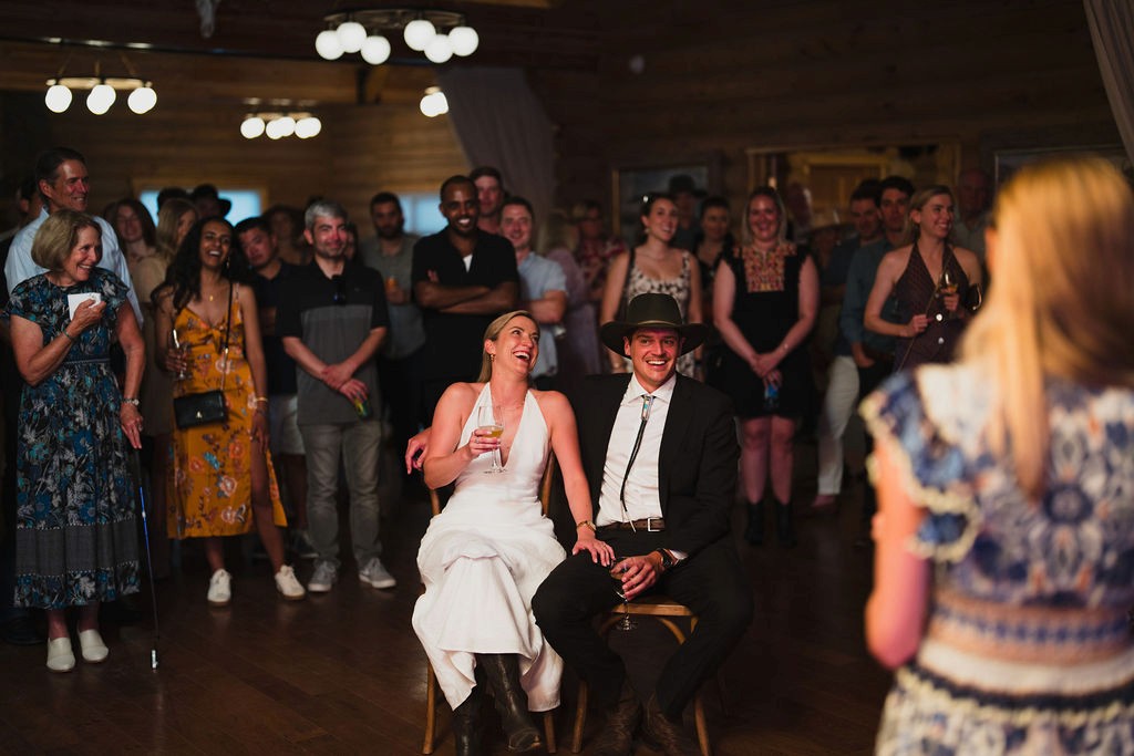 A bride and groom in Western-inspired wedding attire laugh while listening to a reception toast as guests look on.