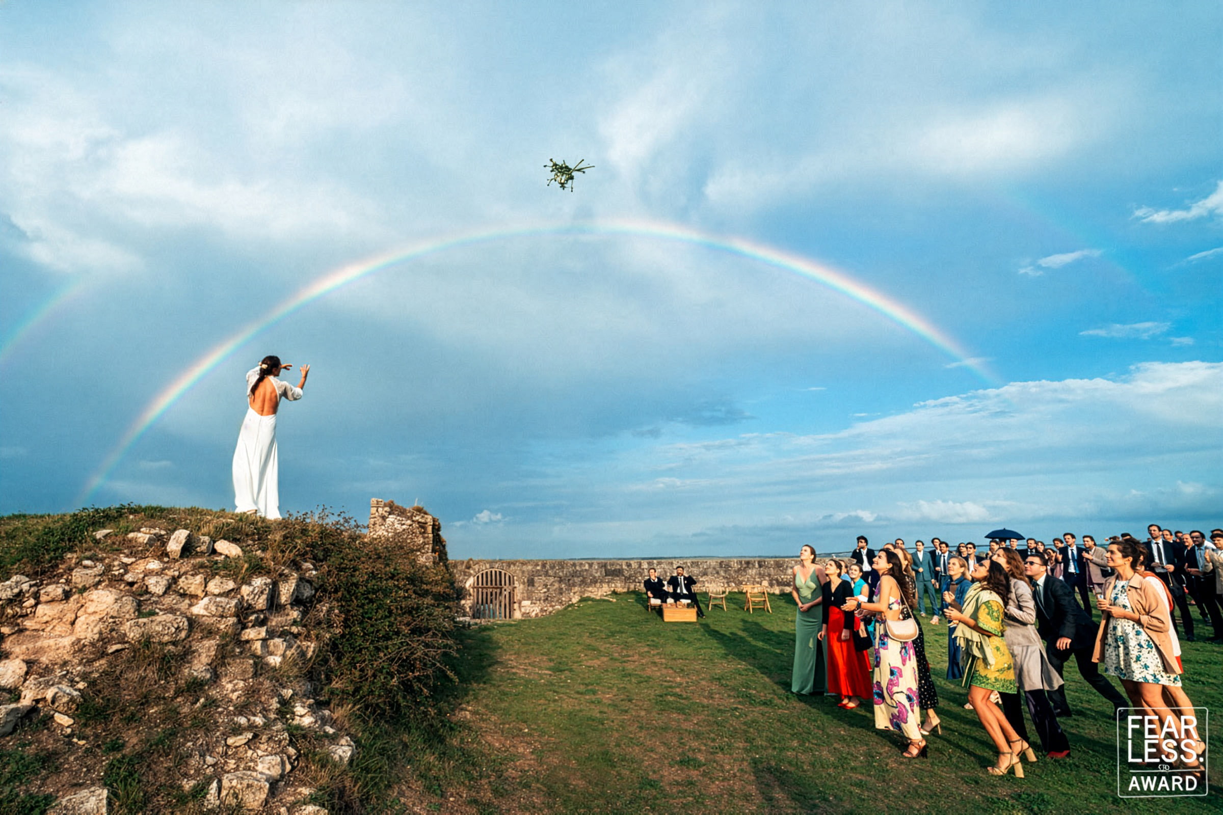 Mariée lançant son bouquet depuis les remparts du fort La Prée sur l’île de Ré, sous un arc-en-ciel spectaculaire, entourée de ses invités dans un moment de célébration intense et spontané.