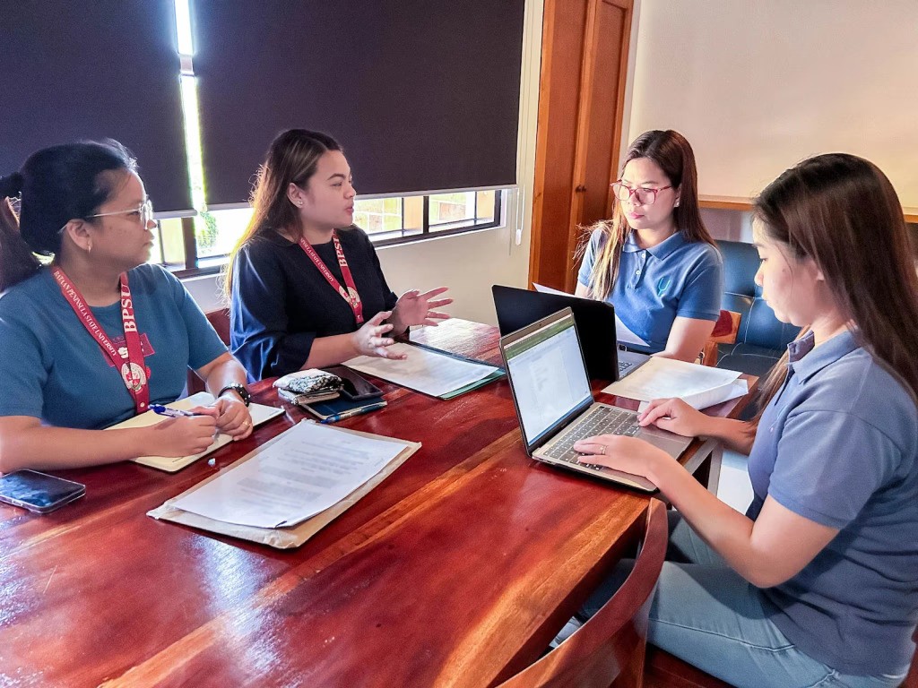 Four women sit around a wooden table in discussion, using laptops and notebooks in a meeting room.