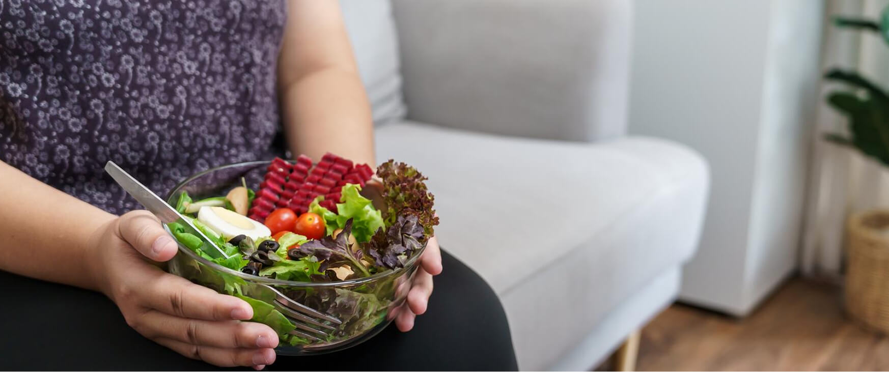 A person sitting on a sofa holds a bowl filled with a colorful assortment of fresh vegetables, including leafy greens, cherry tomatoes, cucumbers, and what appears to be beetroot sticks, reflecting a nutritious postpartum diet inspired by Ayurveda.