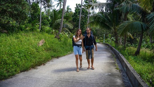 A couple walks hand-in-hand down a scenic road surrounded by lush greenery.
