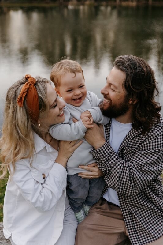 Husband and wife holding their young child during family photoshoot beside a pond in Jeffreys Bay
