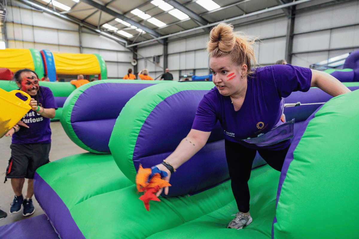 Outdoor team-building event with participants wearing matching blue shirts, taking part in inflatable obstacle courses and games, smiling and cheering, with the words 'ENTER A TEAM' in large bold white and green text across the image.
