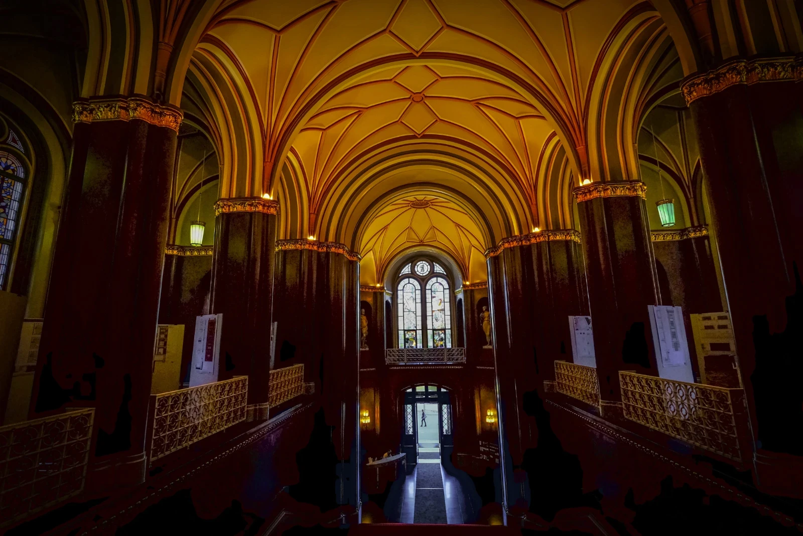 Ornate Gothic-style hallway with high, arched ceilings, illuminated by soft, amber lights. Stained glass window and intricate detailing create a grand ambiance.