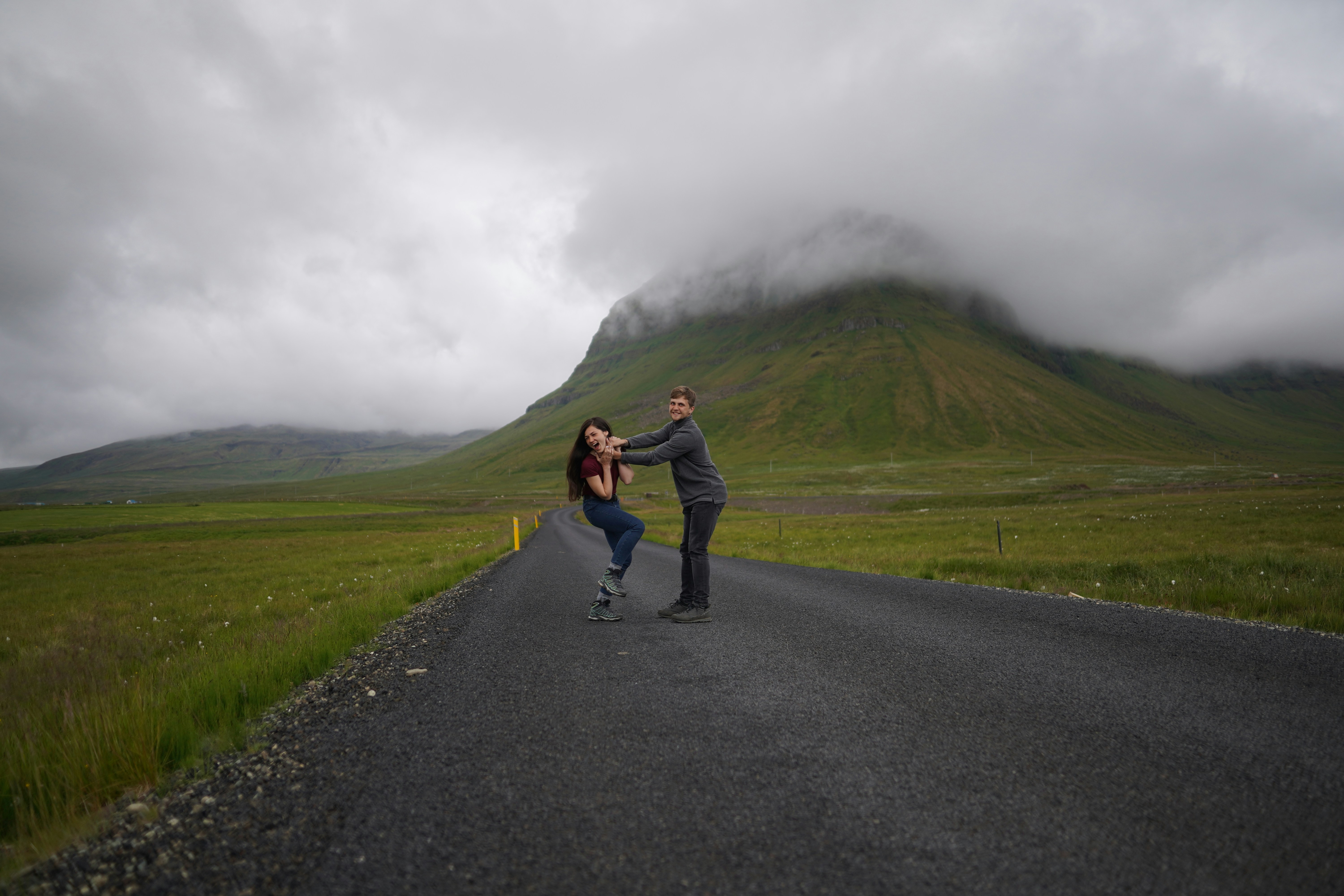 a man and woman sharing a kiss on a road with grassy hills rolling behind them