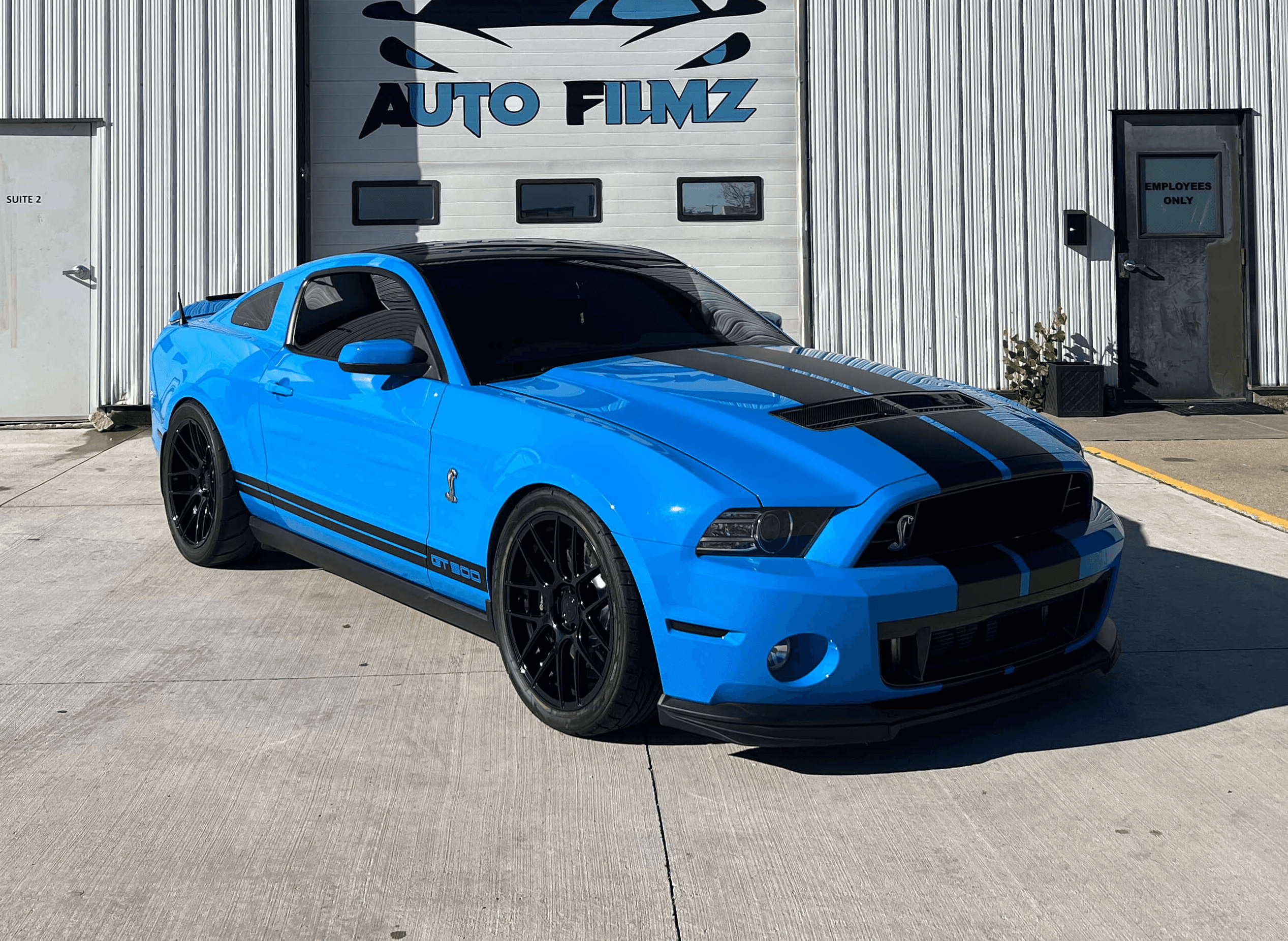 A vibrant blue Ford Mustang Shelby GT500 with black racing stripes is parked on a concrete driveway in front of a white industrial building.