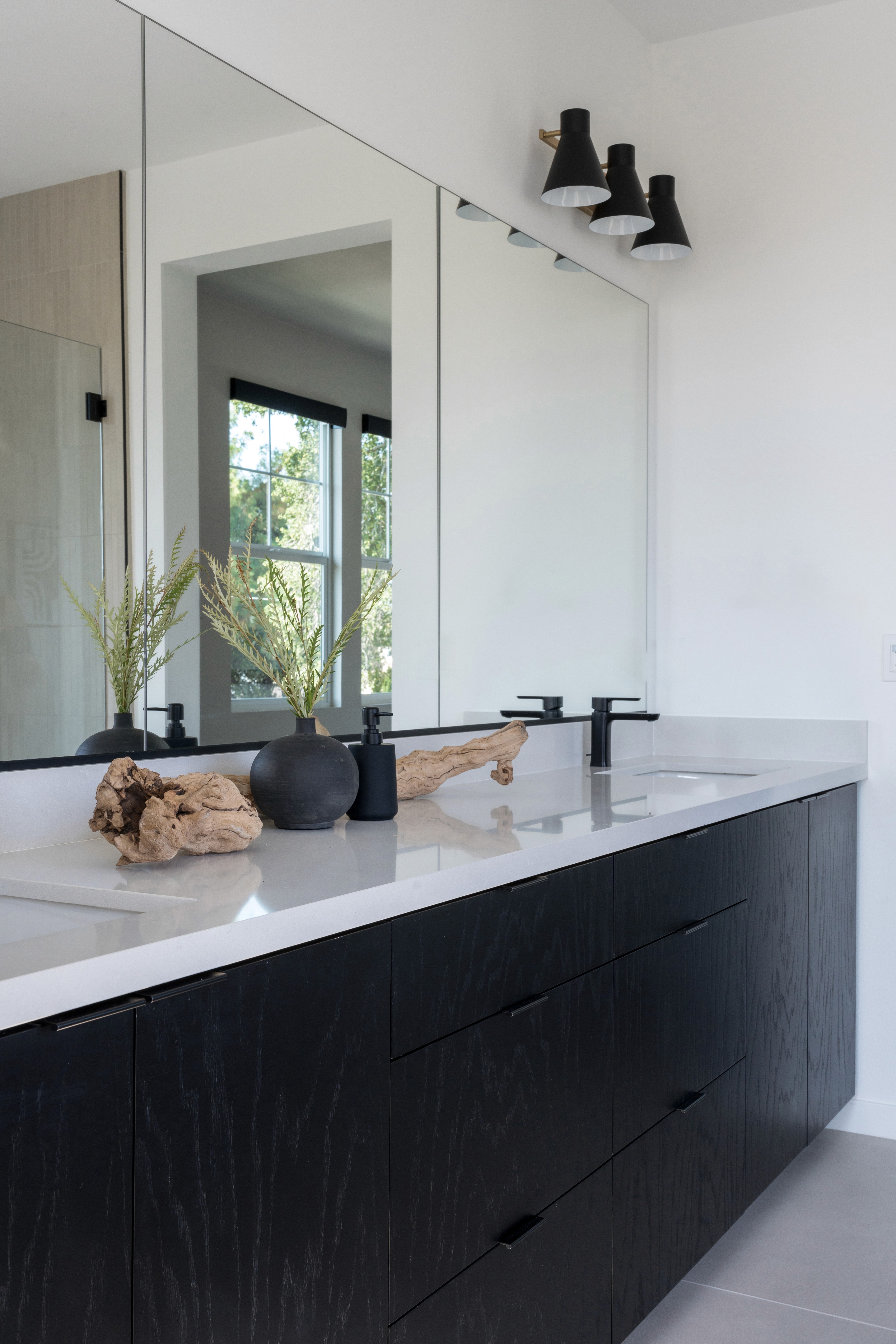Primary bathroom with neutral wood floor, black wood cabinetry, white countertops, vanity lighting, dual sinks with black faucets, decorative light wood accents, and black vases  by Jubilee Interiors in Arcadia, California