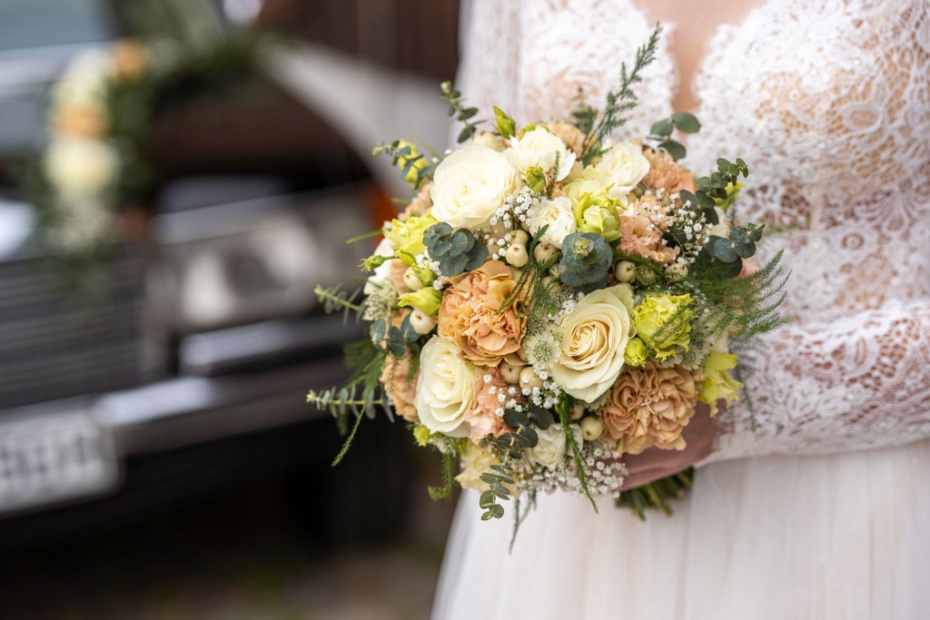 Braut hält Brautstrauß mit hellen Blüten vor Oldtimer während Hochzeit
