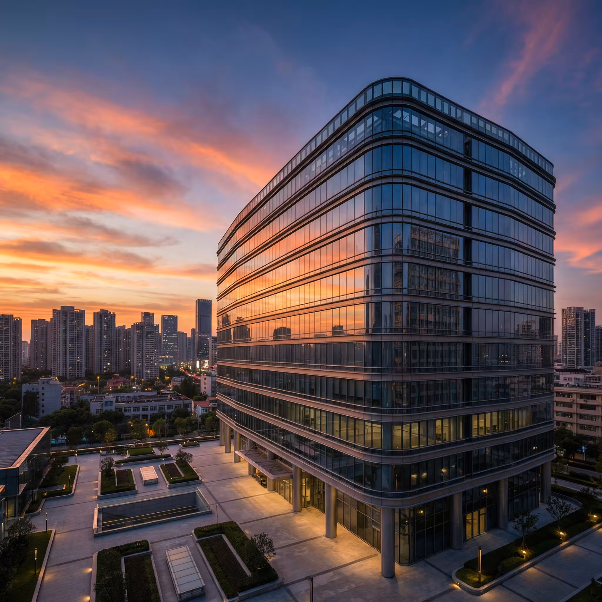 Modern glass office building photographed from below against a colorful sunset sky with city skyline behind.