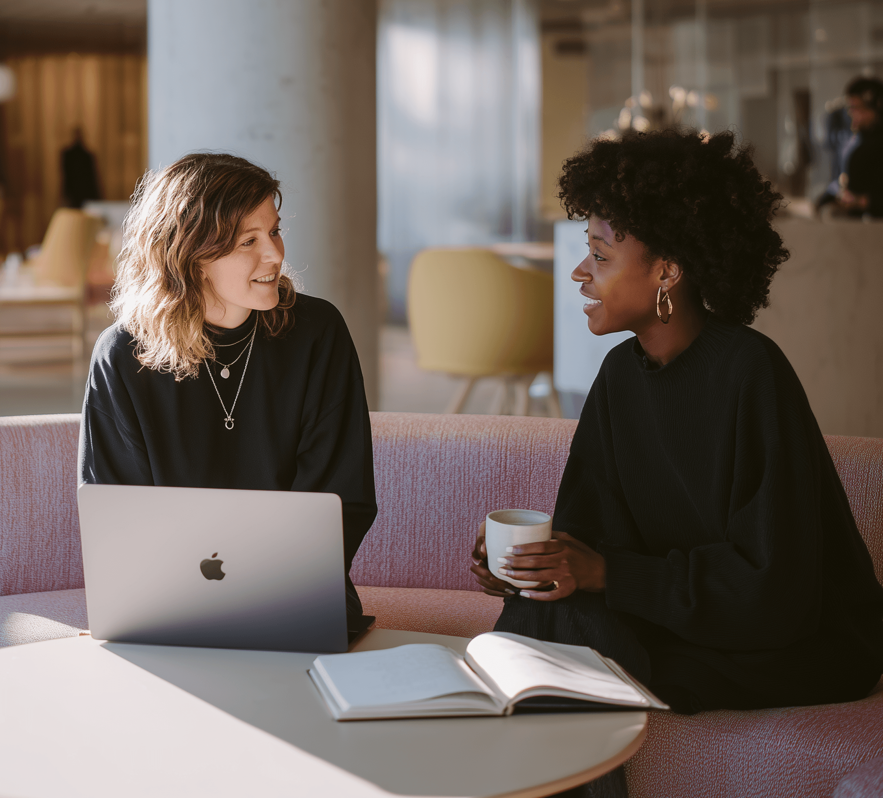A trendy and cozy work environment where two people chat over a laptop and notebook.