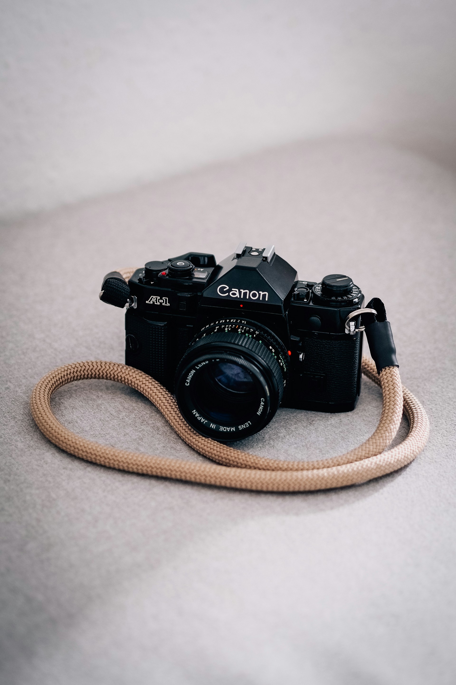 Black Canon film camera with a tan wrist strap resting on a light gray surface.