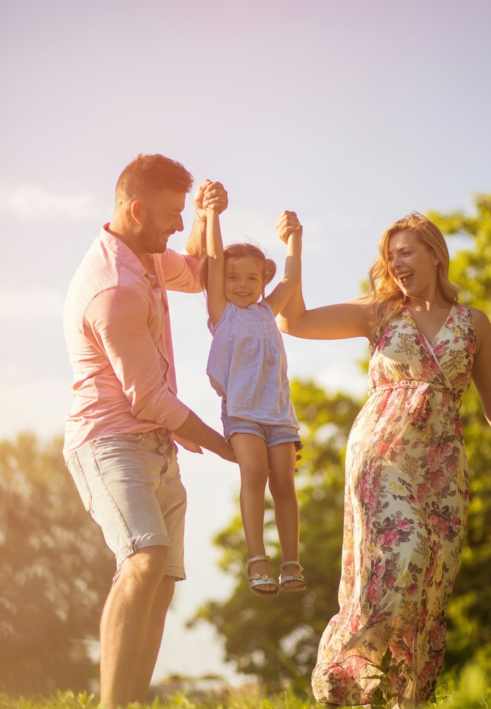 A joyful family enjoys a sunny day outdoors, with the parents lifting their smiling daughter by the hands, creating a heartwarming scene of love and togetherness in a natural setting.