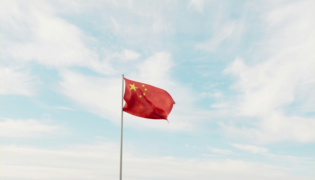 The Chinese national flag, a red field with five yellow stars, flutters against a bright blue sky with scattered white clouds, symbolizing China's identity and national pride.