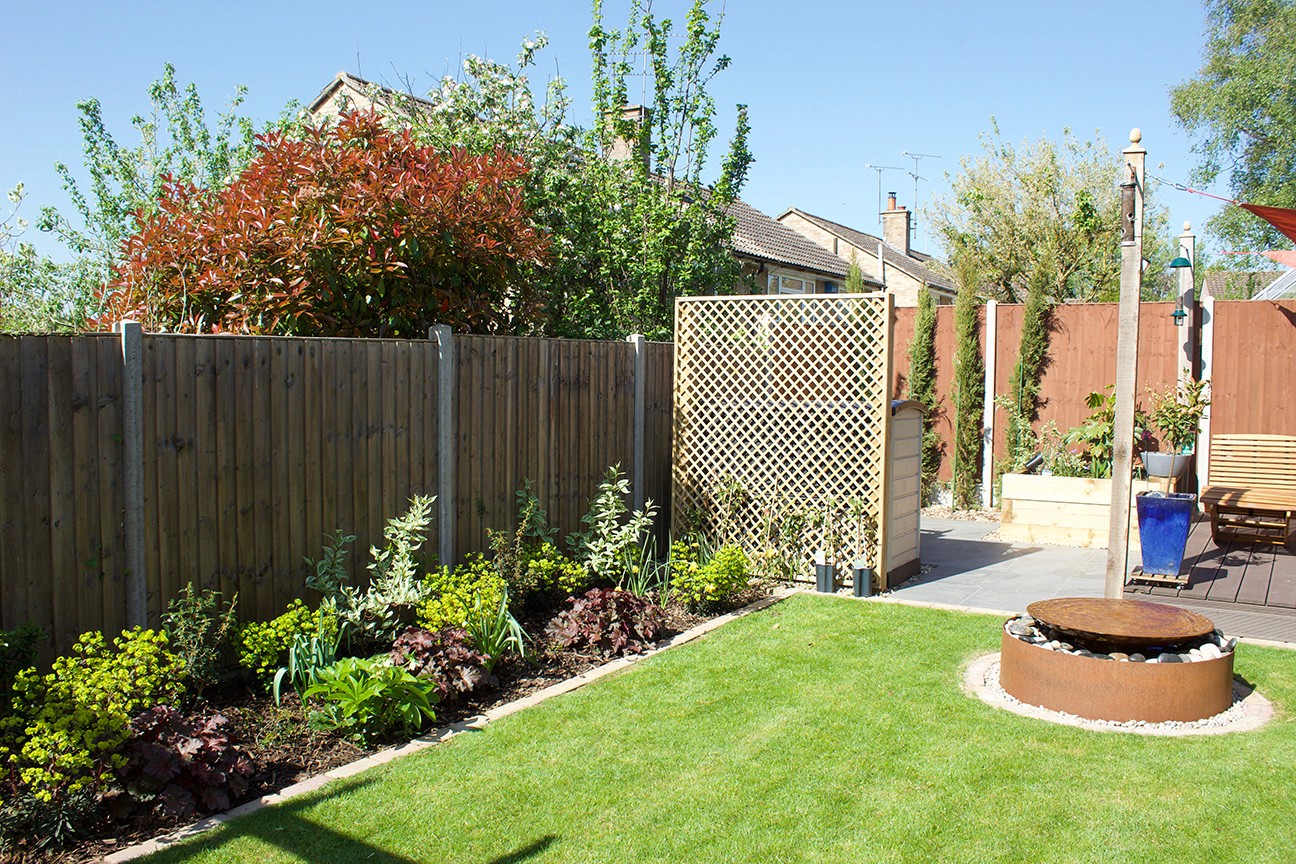 A sunny garden with green grass, landscaped plants, a wooden fence, and a circular stone fire pit.