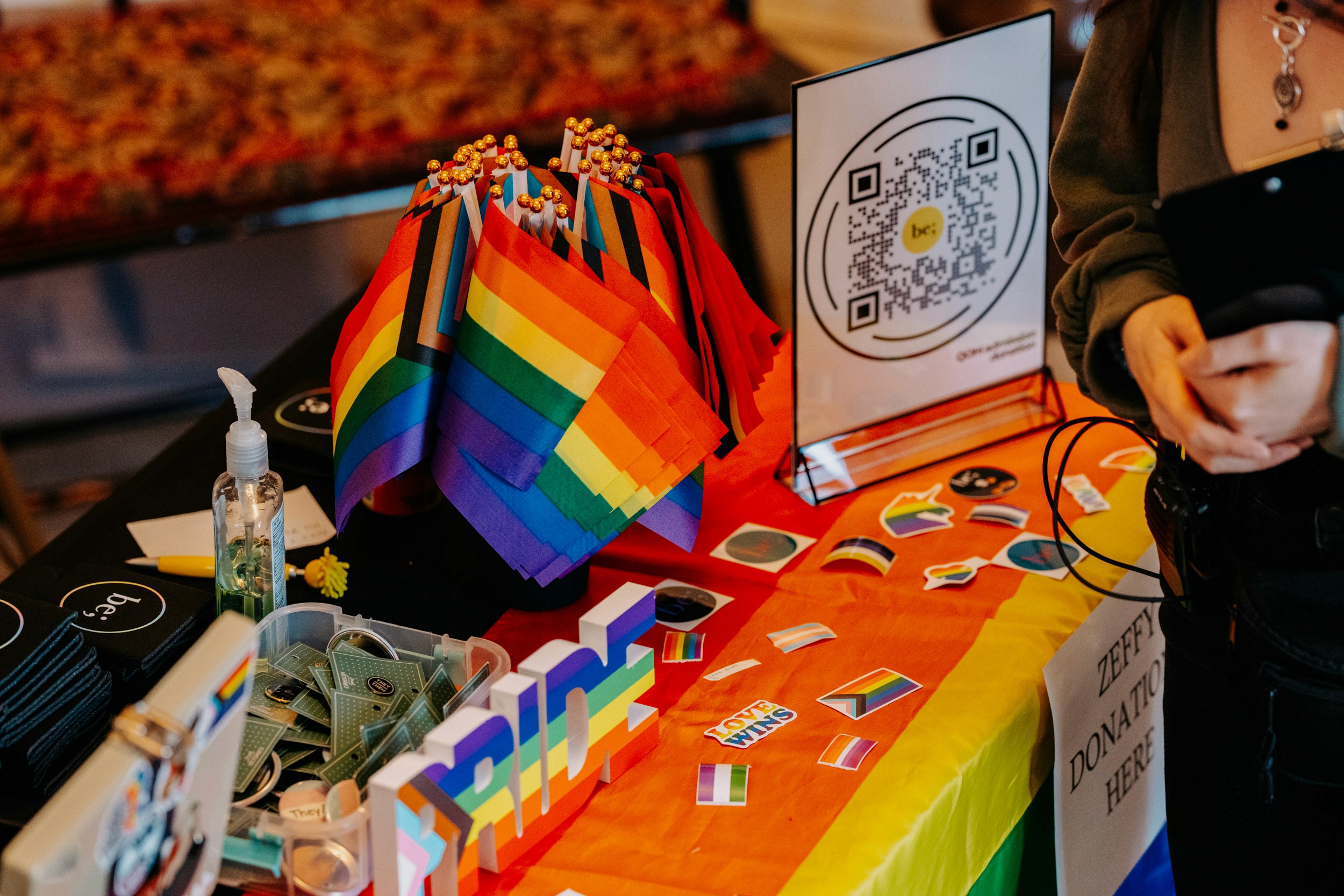 Pride flags, stickers, and donation table setup at the Queer Open Mic (QOM) hosted by be; community in Bridgewater, Massachusetts