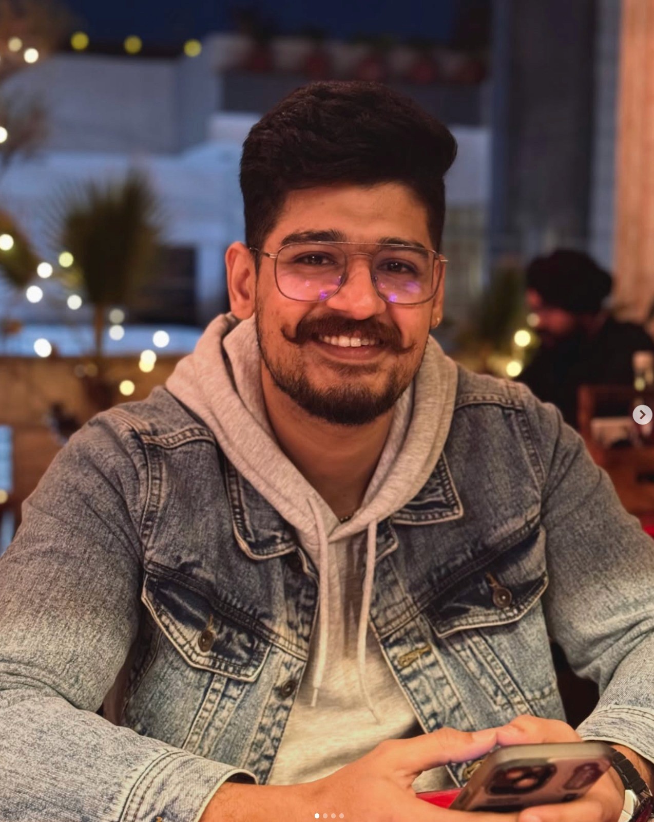 Young man with wavy hair wears a dark overcoat, rust-colored sweater, white shirt, and patterned tie, exuding a poised and sophisticated demeanor.