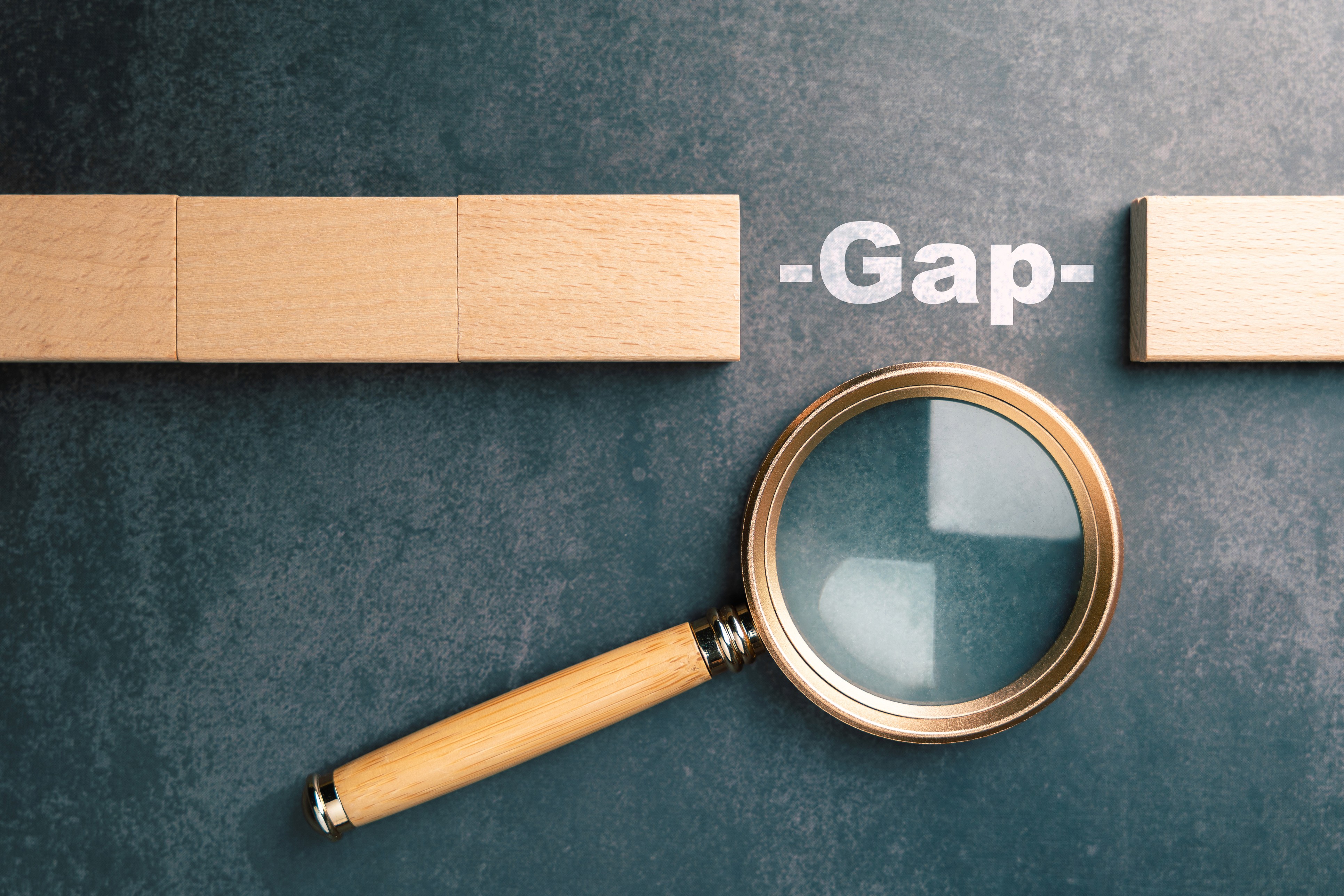 Magnifying glass focusing on the word gap between wooden blocks representing gap analysis, business strategy, performance evaluation, improvement planning