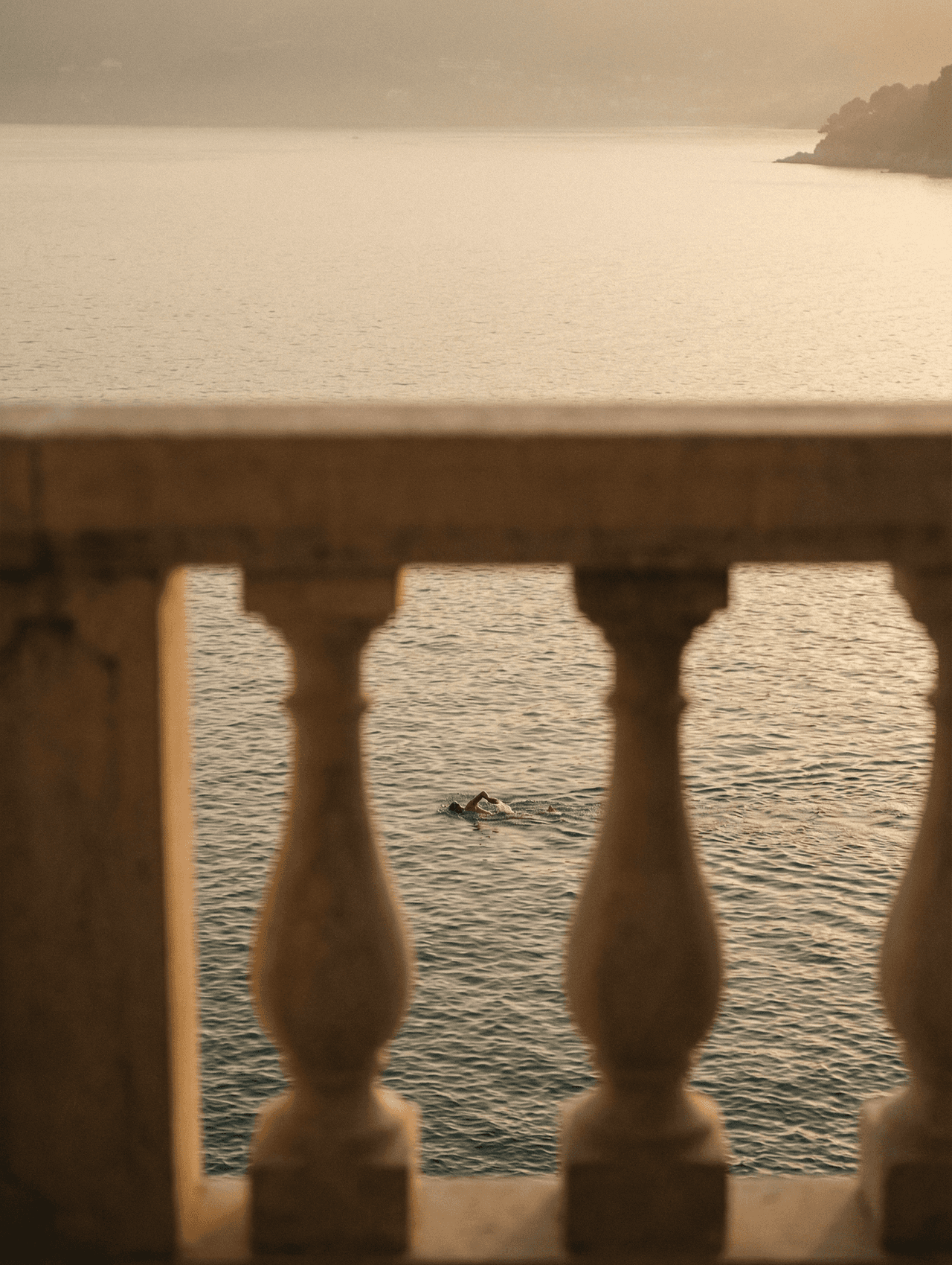 Calm sea at golden hour seen through a stone balustrade, with a lone swimmer in the distance.