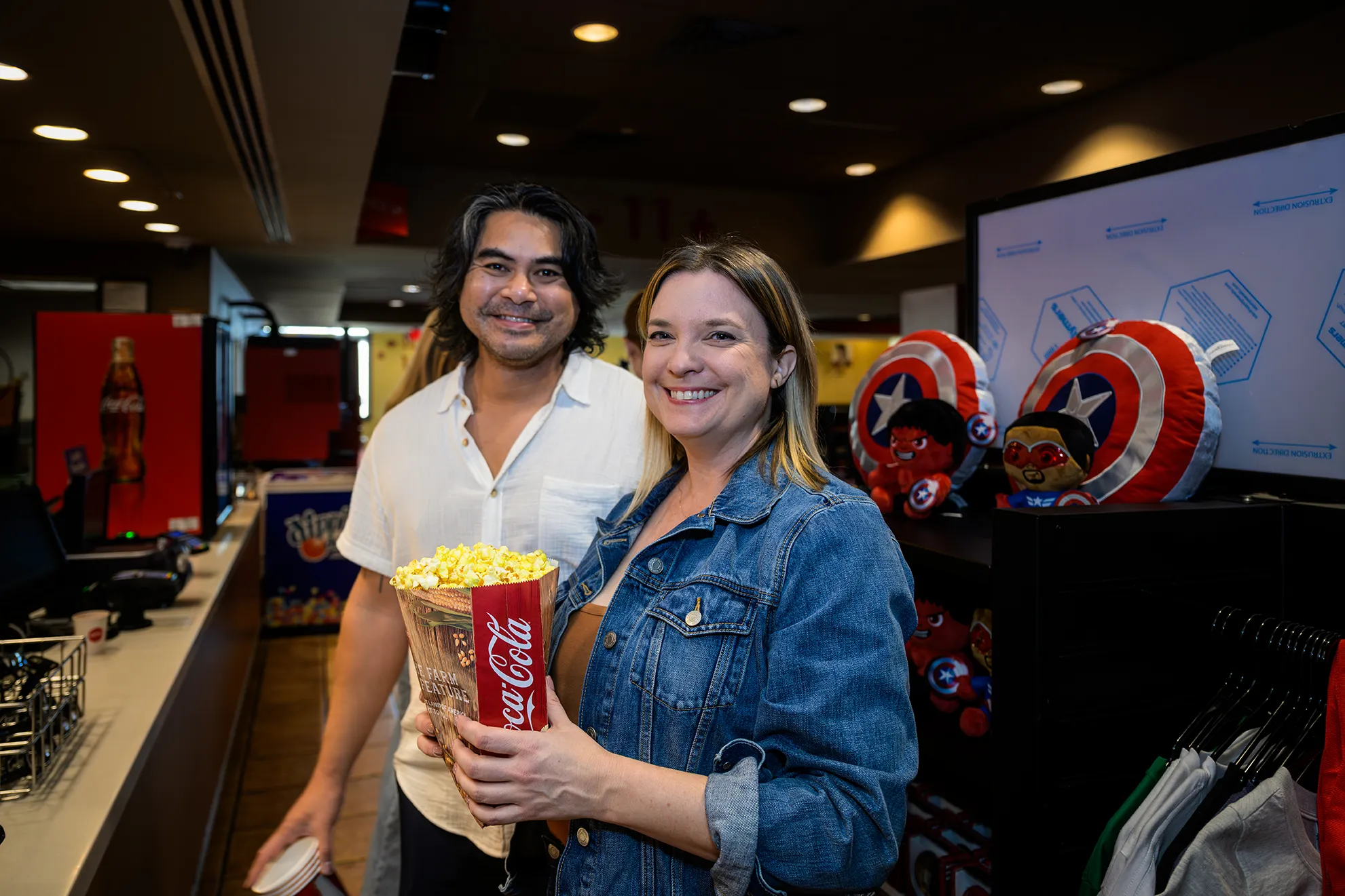 Couple holding a Coca-Cola popcorn tub in the AMC Alamo IMAX lobby, with Marvel merchandise displayed behind them