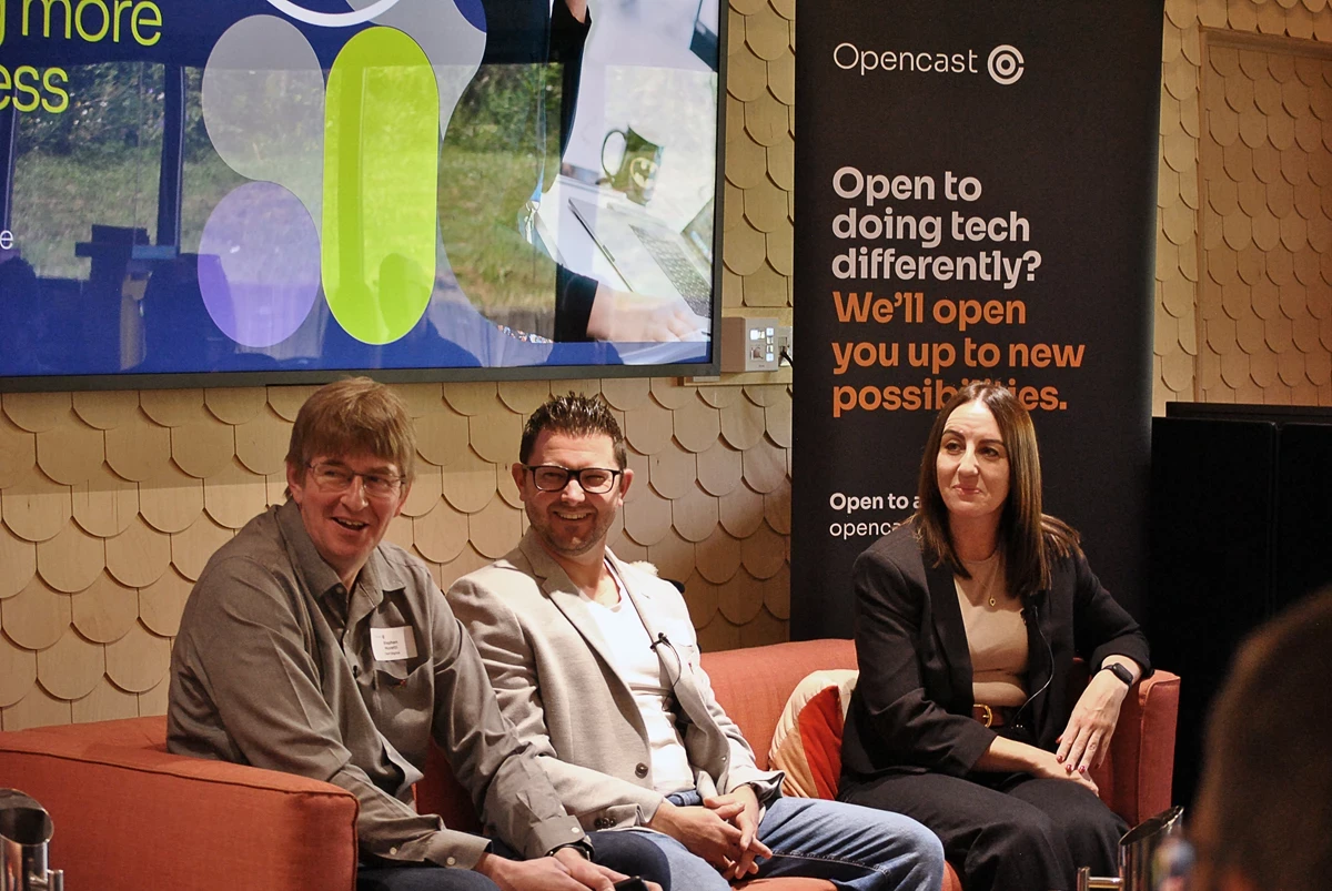 Three people seated on a sofa during a tech event, with an Opencast banner and a large screen in the background displaying graphics and text.