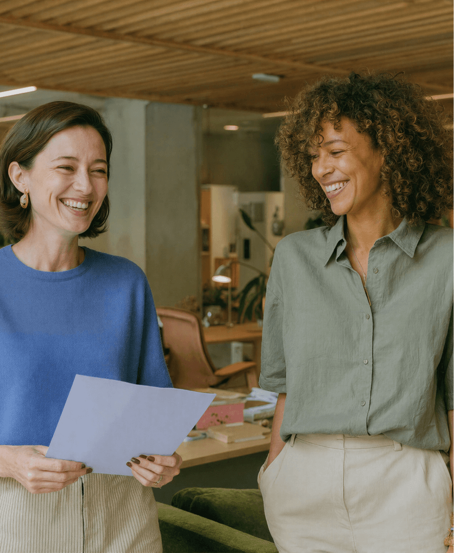 Duas mulheres sorrindo enquanto analisam um documento juntas em um escritório moderno cercado de plantas, representando a comunicação eficiente entre profissionais de clínicas com o suporte da Tivita.