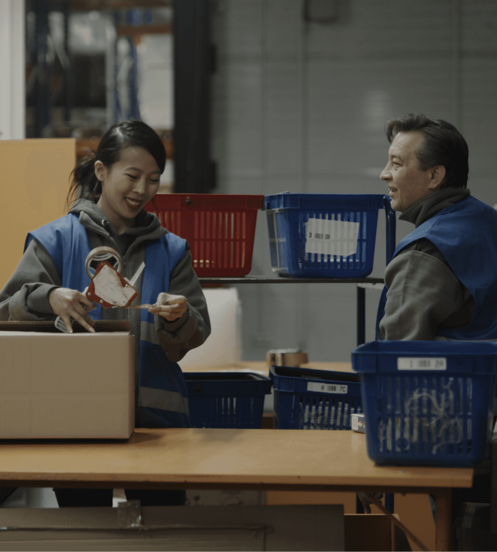 Two warehouse workers in blue vests chatting and smiling at a workstation surrounded by bins and boxes