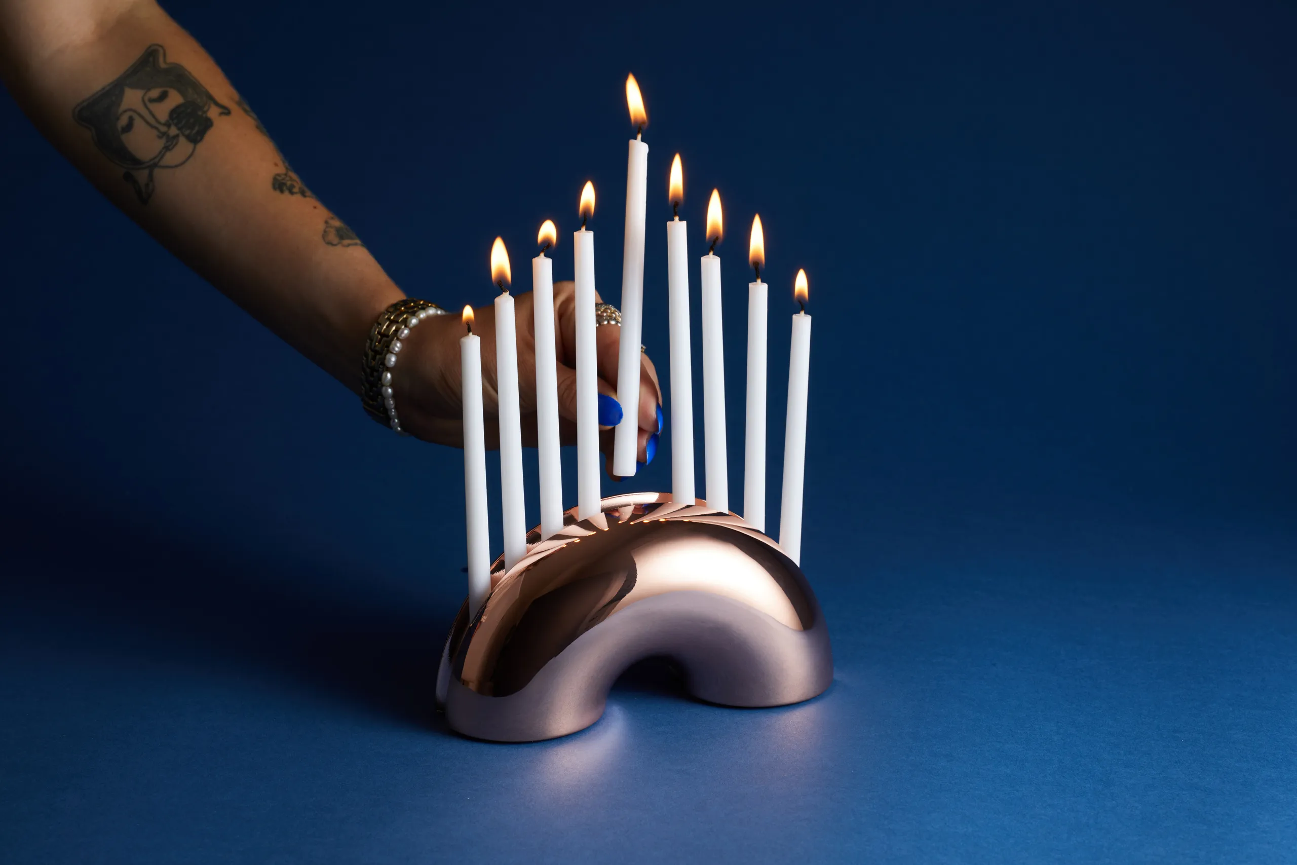 Hand reaching toward the lit metallic Nosh Menorah on a dark blue background