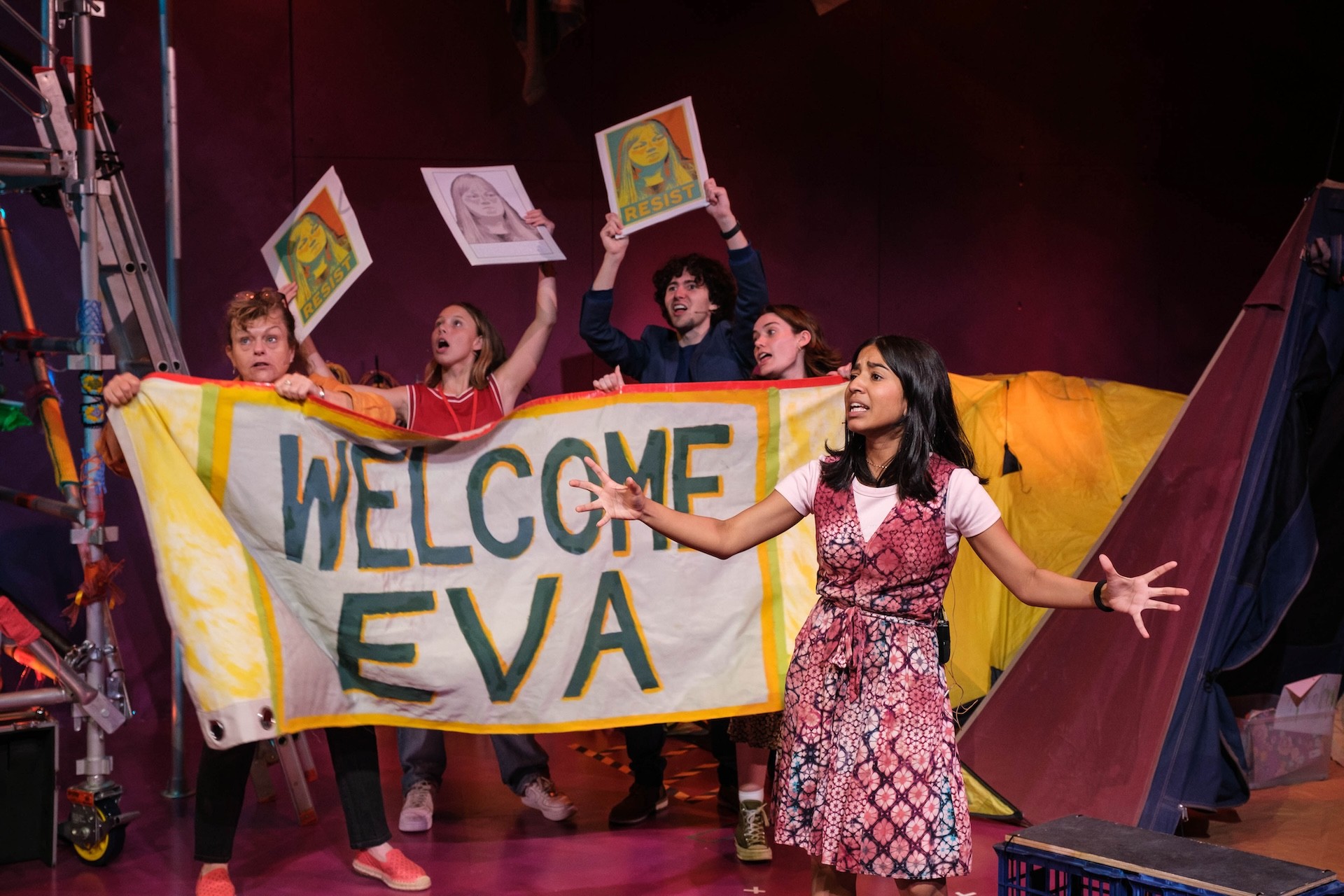 A group of young activists stand behind a large hand-painted banner reading “WELCOME EVA,” holding protest-style posters above their heads. In front, a performer in a patterned dress speaks passionately with outstretched arms on a brightly coloured stage.